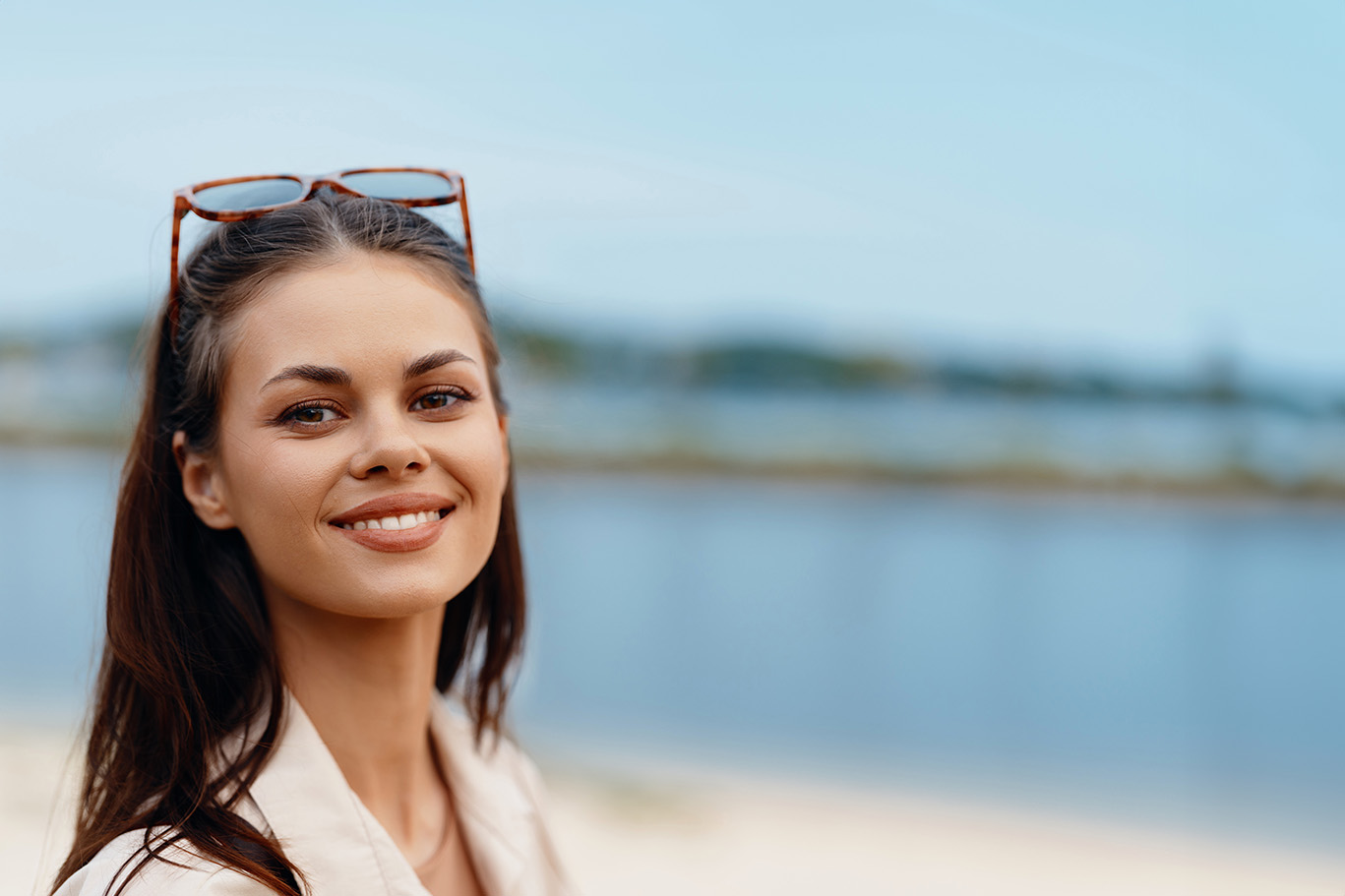 A woman with a radiant smile, wearing sunglasses and a white top, stands against a backdrop of a body of water and a clear sky.